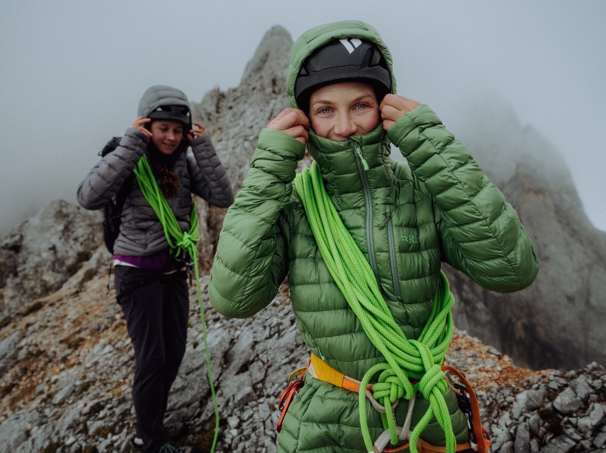 A women in a green jacket pulls up her hood. Another woman behind her has a rope slung over her shoulders.
