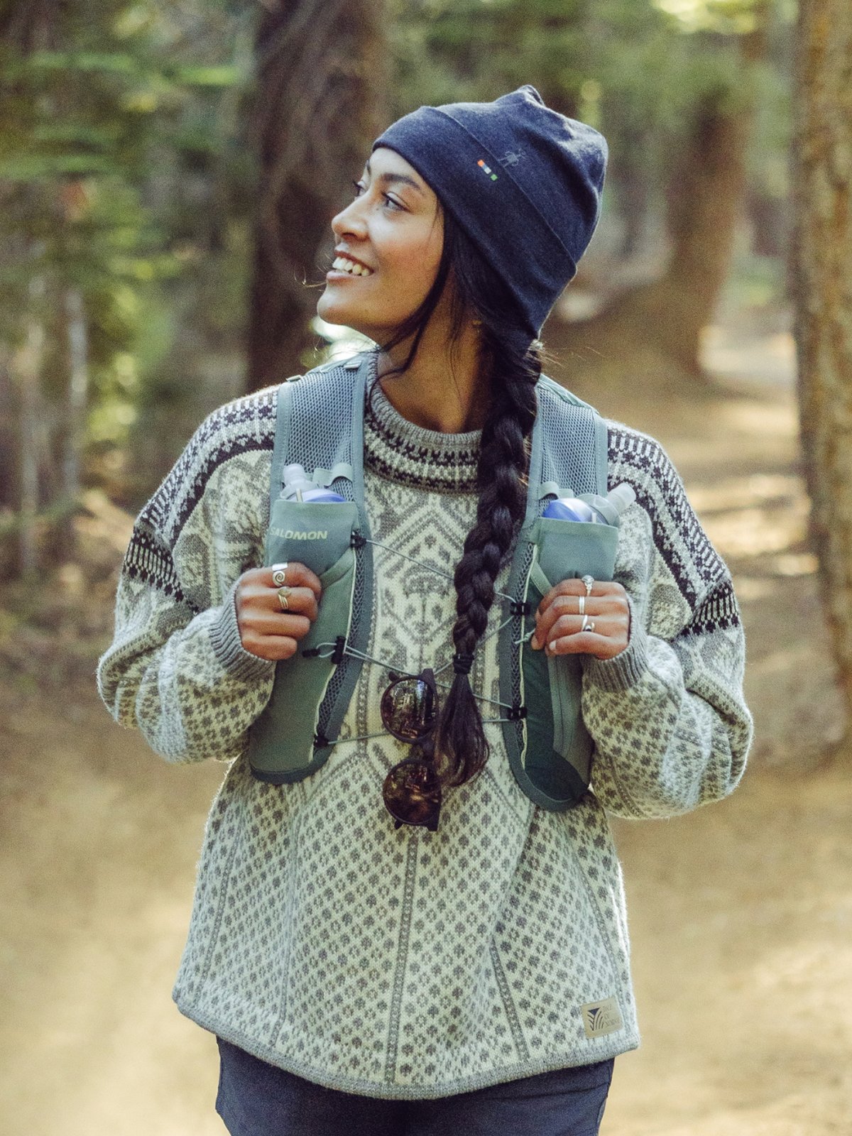 A person with a long braid and a navy beanie walks on a forest trail, wearing a patterned knit sweater and carrying a hiking vest with water bottles and sunglasses attached.