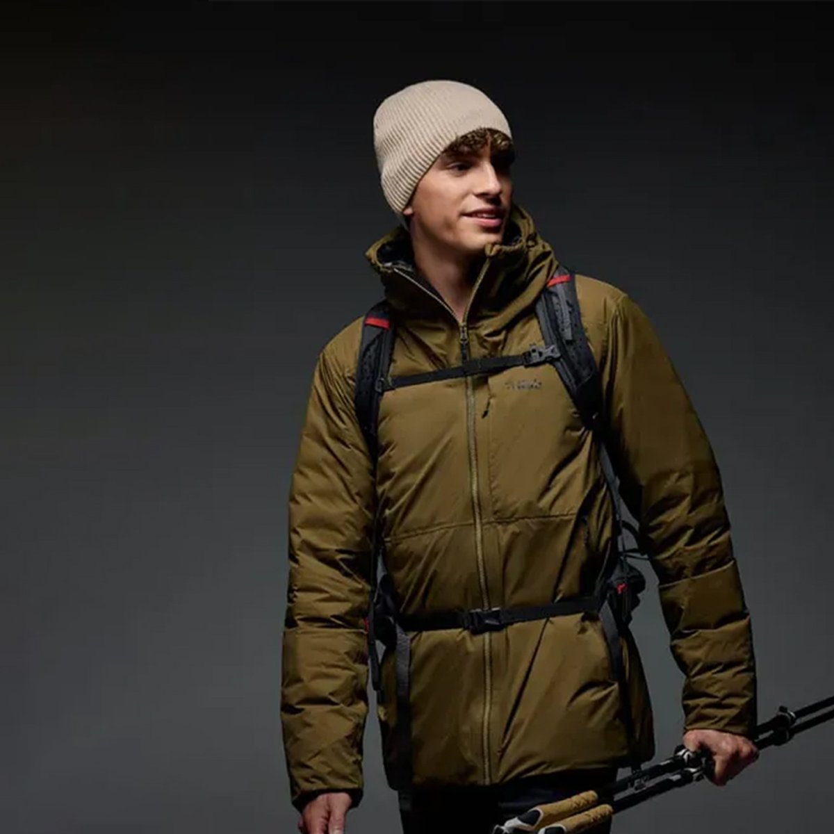 A man wears a hiking pack, beanie, and Rab Microlight jacket. He holds hiking poles in one hand.