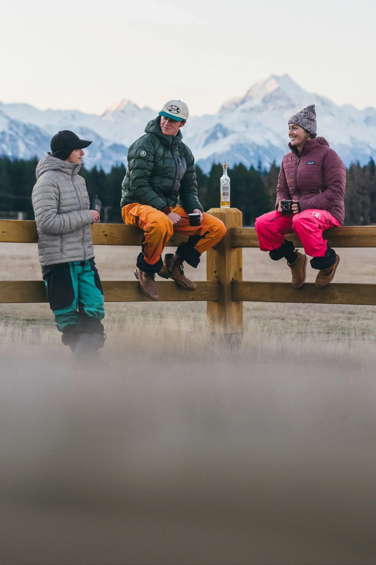 Three friends in insulated jackets and ski pants gather on a wooden fence for après with mugs, snow-covered peaks behind them.