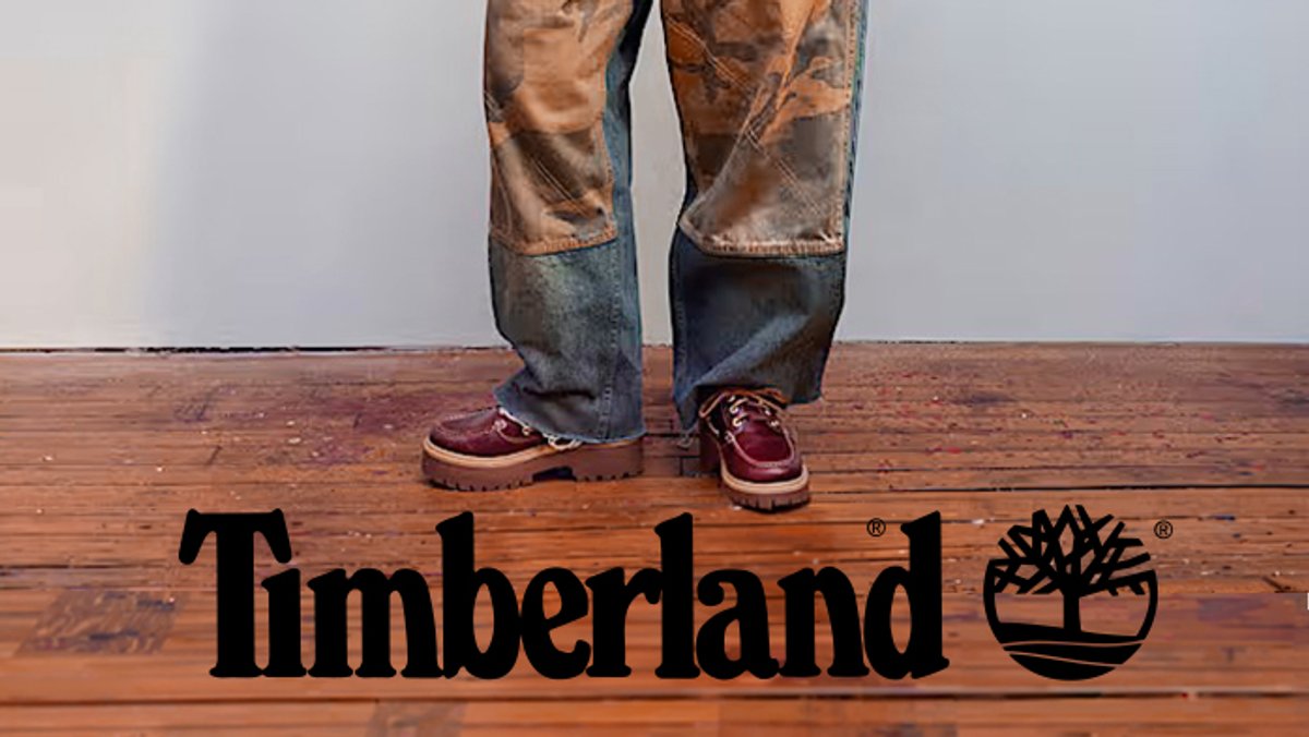 A person wearing faded brown and blue jeans stands on a wooden floor next to the Timberland logo and tree icon, showcasing burgundy waterproof boat shoes.