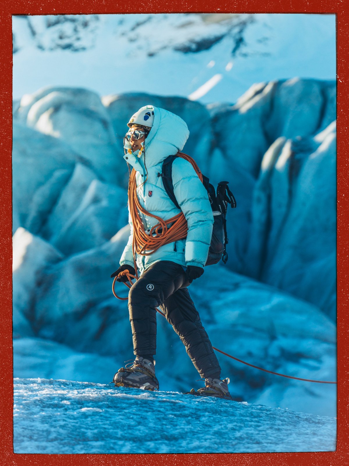 Climber in a helmet and crampons with a coiled rope over one shoulder, walking a glacier with blue ice formations behind.
