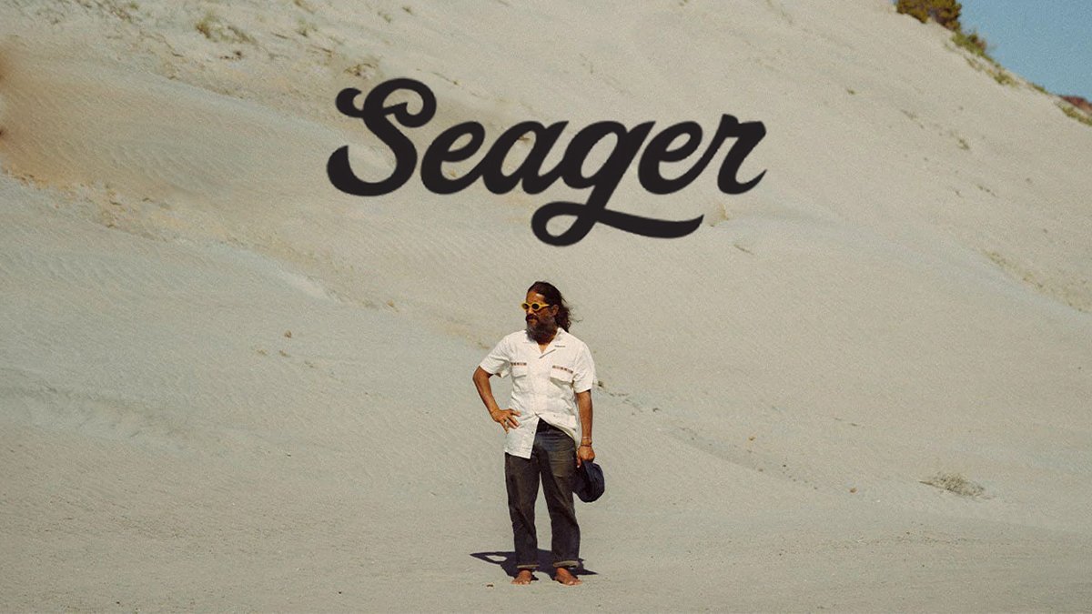A man wearing sunglasses and casual clothing stands confidently against a large sand ridge with the "Seager" logo above him.