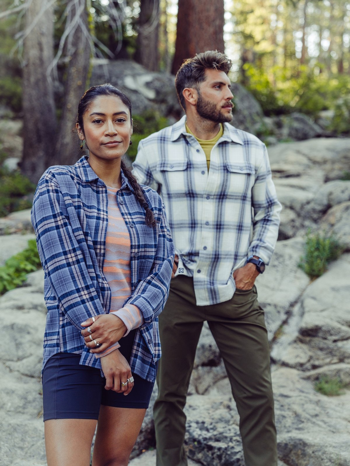 Two people stand outdoors on rocky terrain; one is wearing a blue plaid shirt over a pink-orange striped top and shorts, while the other is dressed in a light plaid shirt and olive green pants.