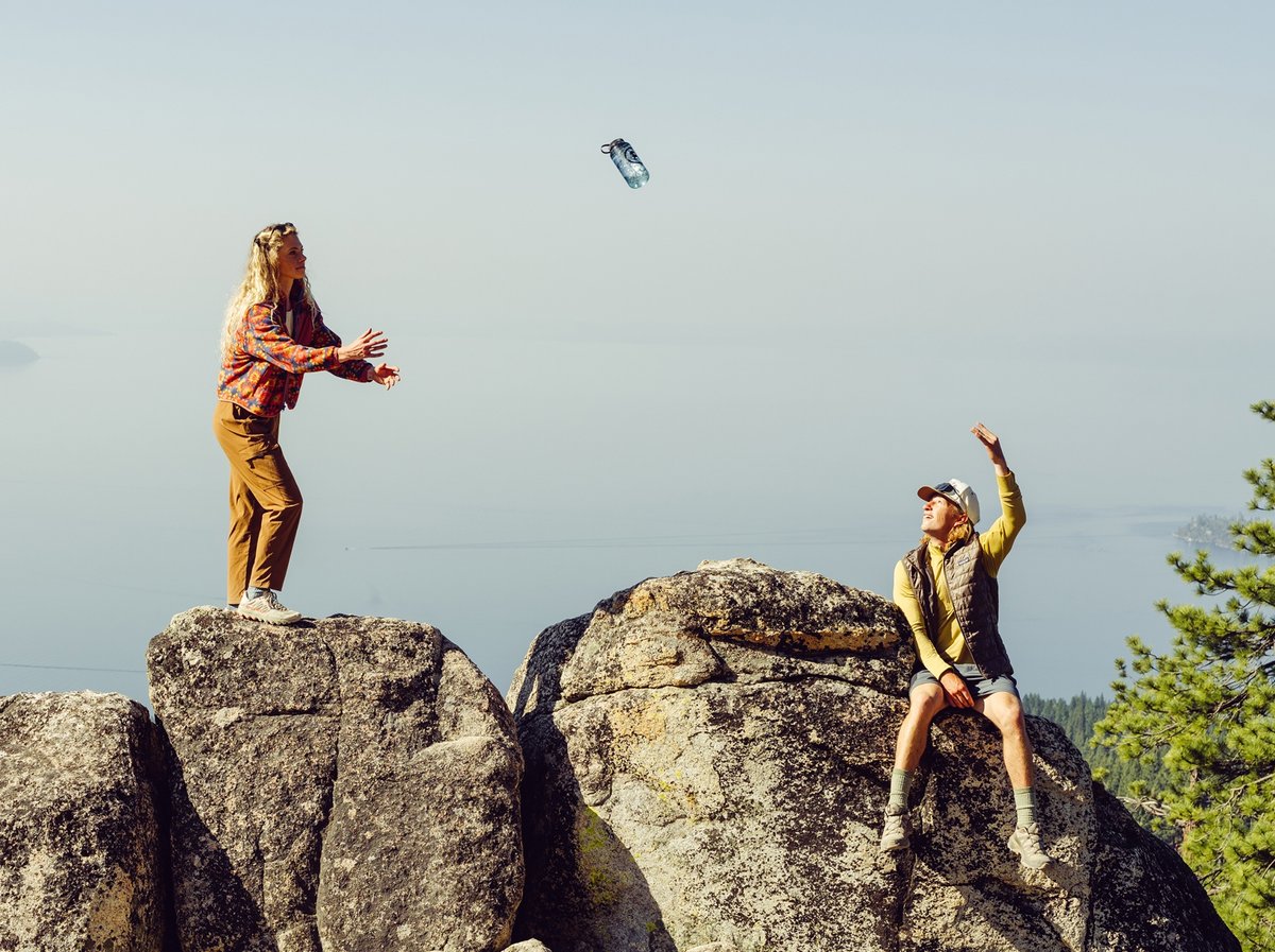 Two hikers toss a water bottle across granite boulders above a hazy lake basin, pine trees framing the ridge behind them.