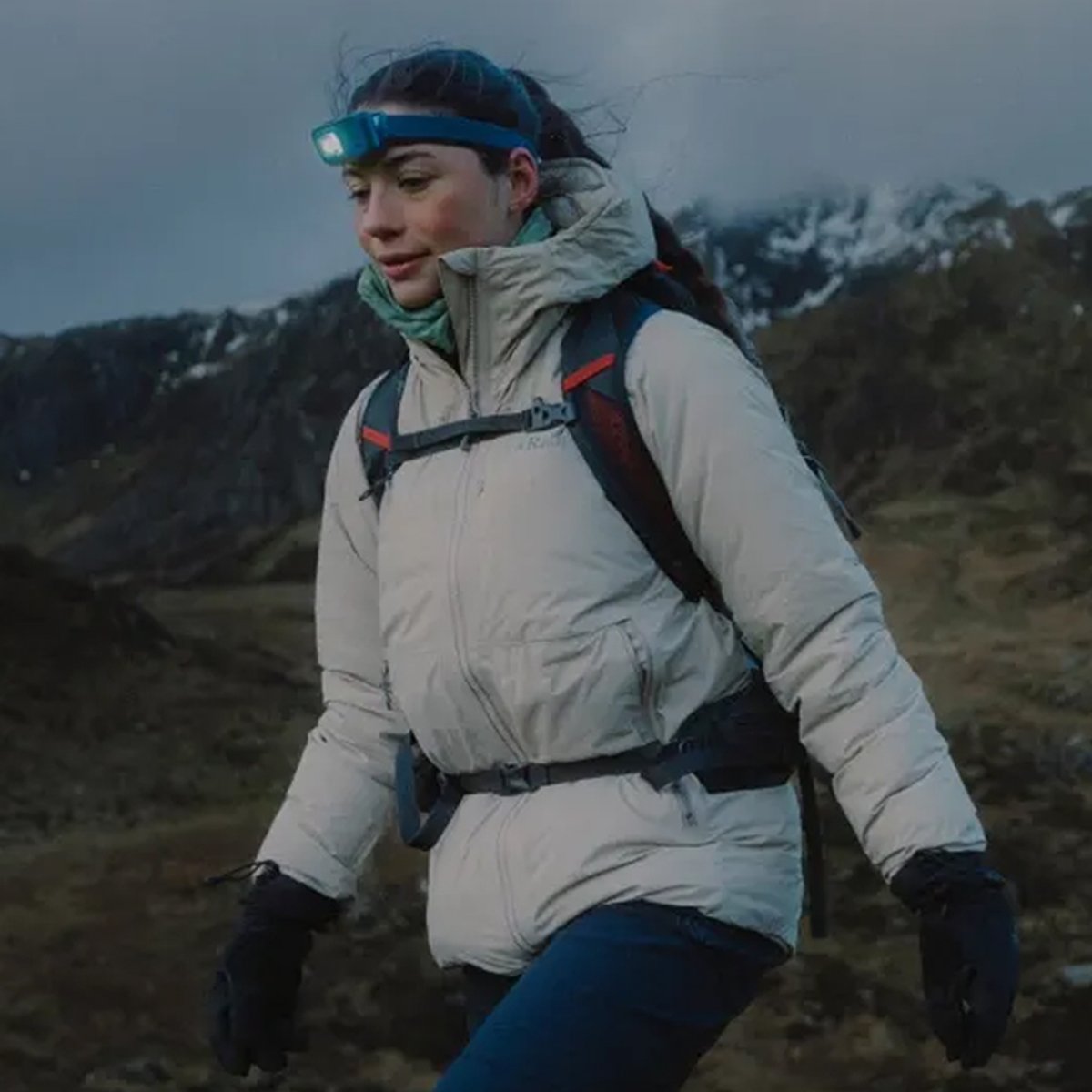 A woman hikes along an alpine ridge, wearing a headlamp, hiking backpack, gloves, and a Rab Microlight jacket.