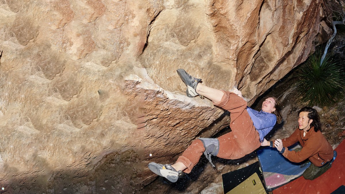 A woman boulders up a rock face, crash pads below her as a man spots her. 