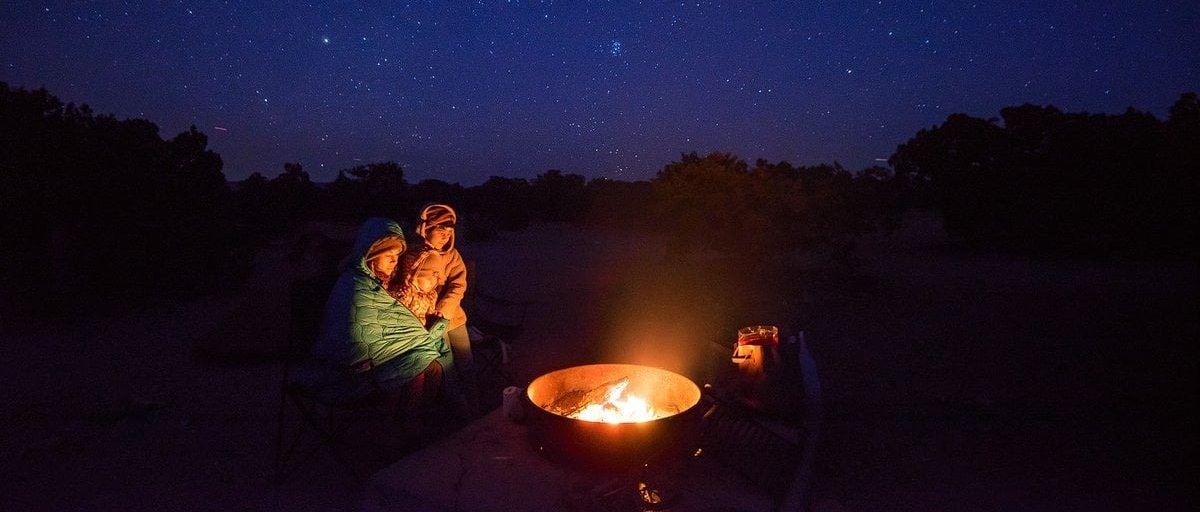 Two campers wrapped in sleeping bags and insulated outerwear sitting beside campfire ring under starry night sky