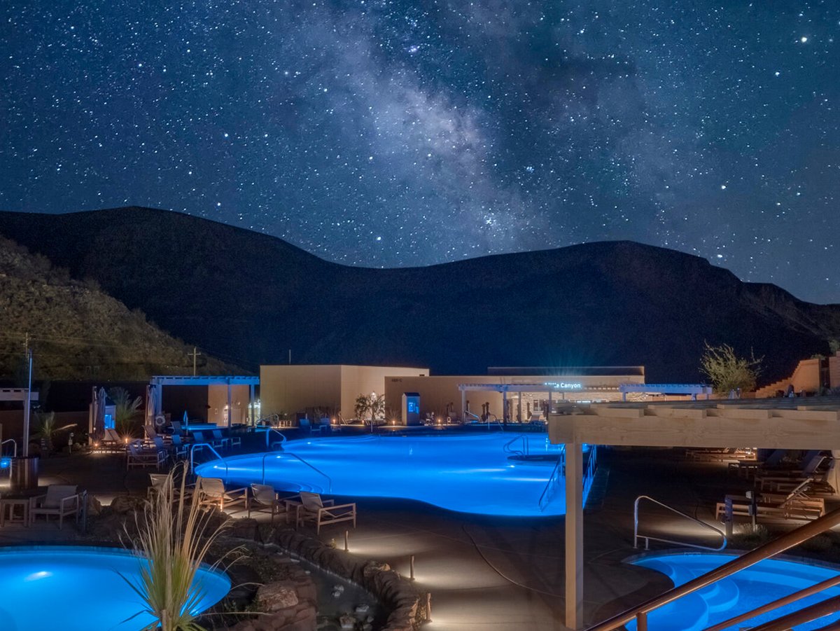 Illuminated outdoor pool complex at night with blue-lit pools, desert landscaping, and canyon ridgeline silhouetted against a Milky Way sky.