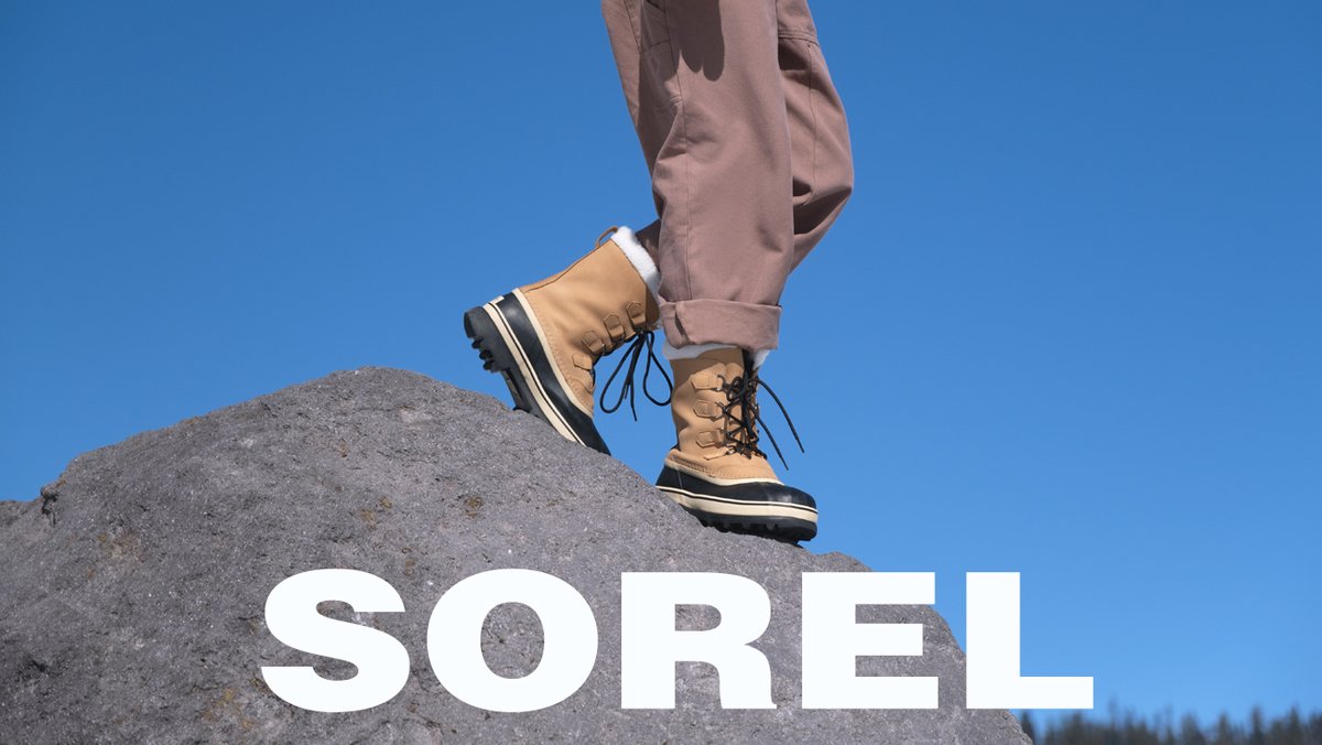 A close-up shows a person wearing tan and black Sorel winter boots standing on a large rock against a clear blue sky.