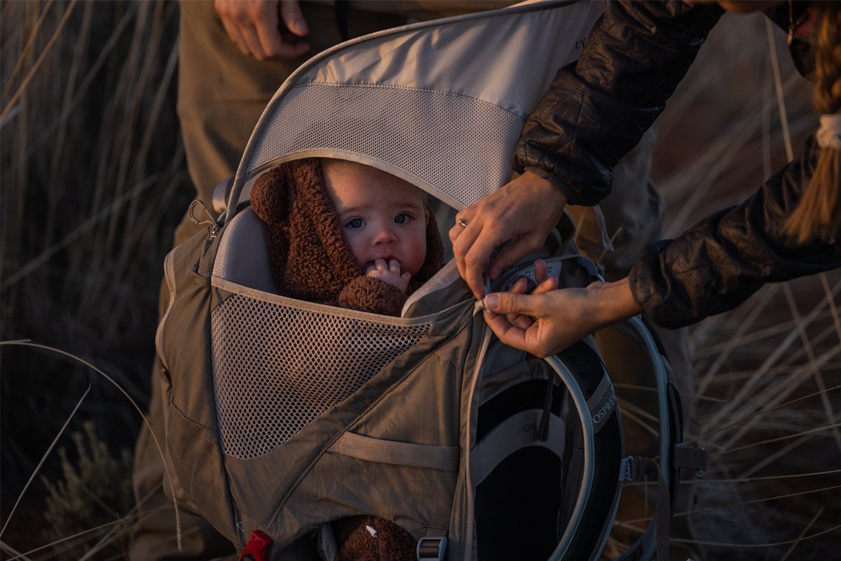 Hands adjust an Osprey child carrier for a baby during sunset.