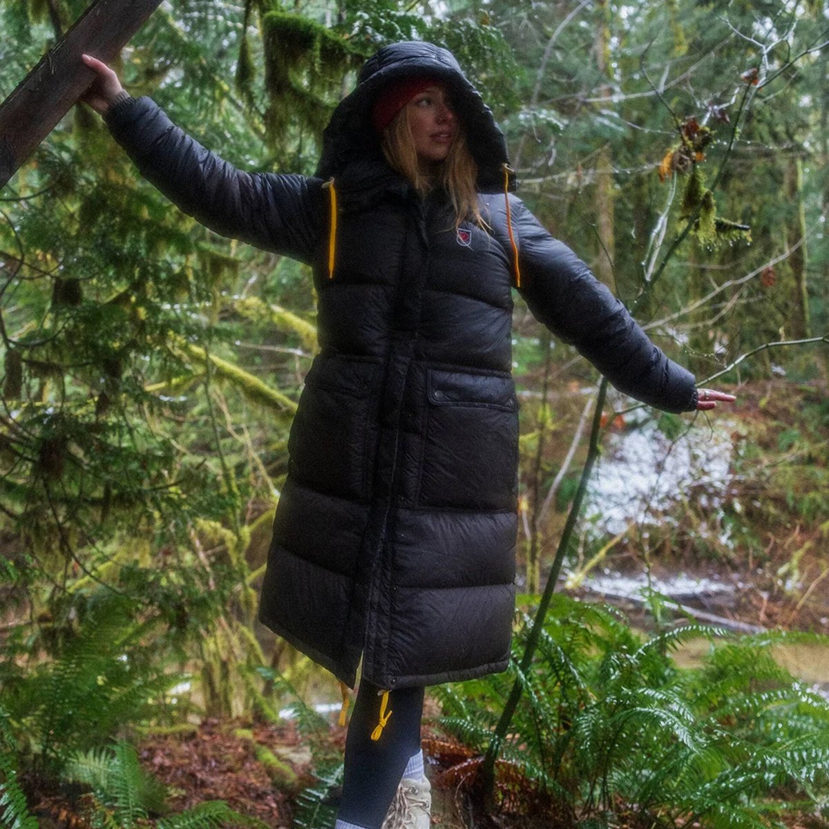 A woman wears a Fjallraven Expedition Long Down Parka against a damp forest background.