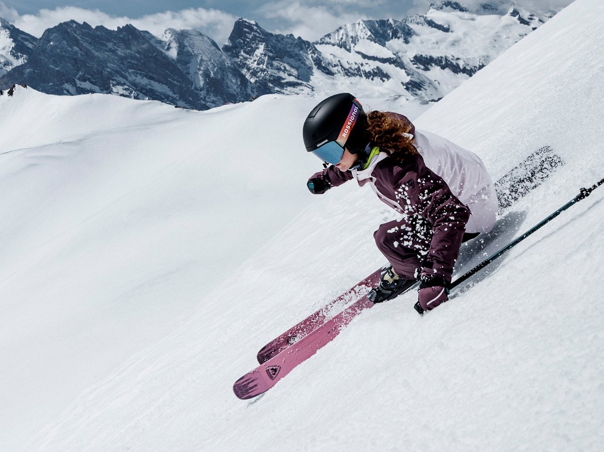 Skier carving a steep, snowy slope in a spray of powder, with jagged snow-capped peaks behind.