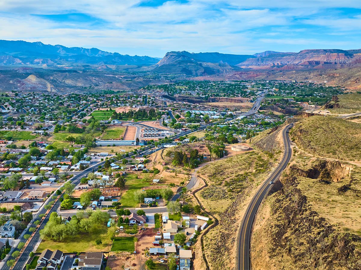 Aerial view of Hurricane, Utah, with a residential street grid spreading across the valley floor, a highway curving along a desert ridge to the right, and red rock mesas in the distance.