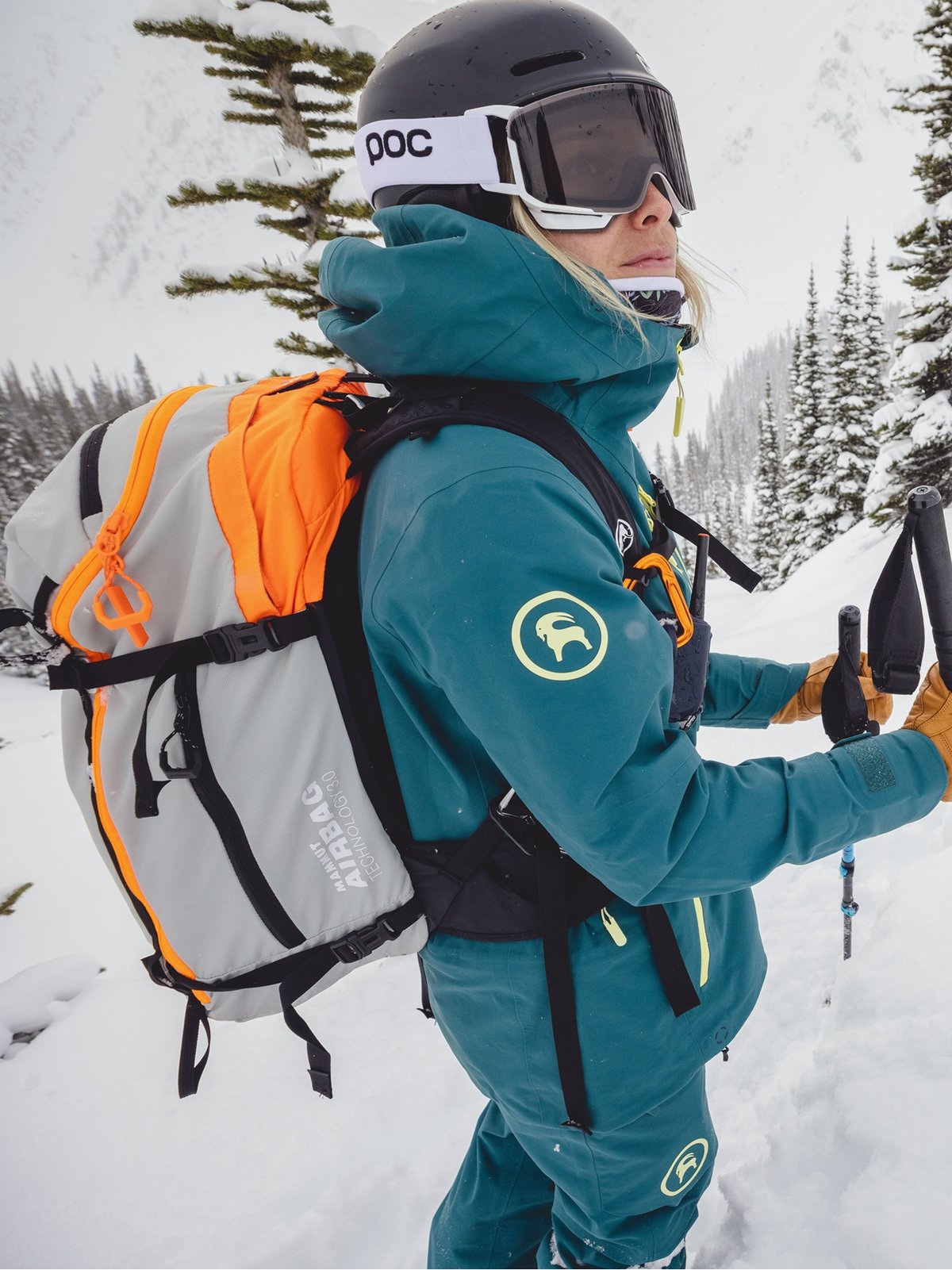 A woman wearing a blue ski and avy bag looks on.