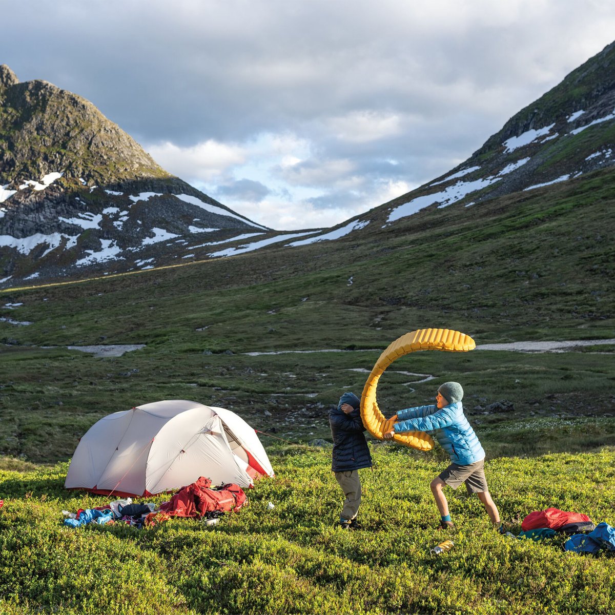 Two adventurers set up a yellow paraglide next to their white camping tent in a scenic alpine valley surrounded by snow-capped mountains and green meadows.