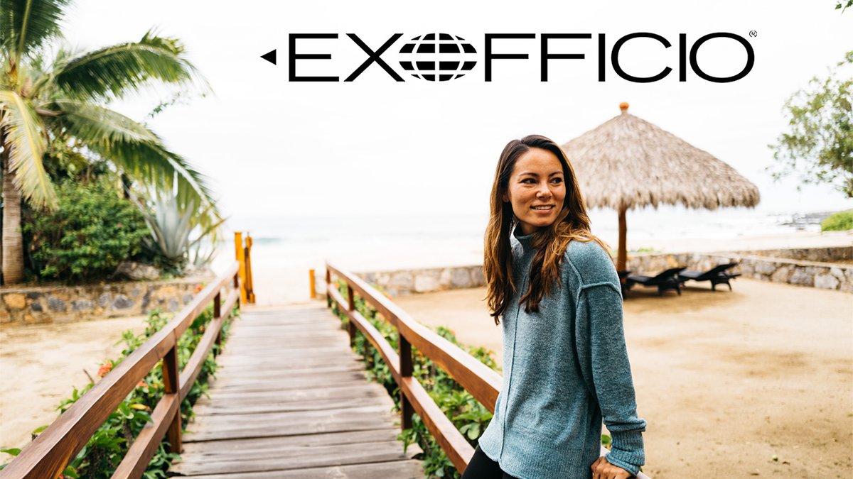 A woman in a blue sweater stands on a wooden beach walkway leading toward the ocean under a centered ExOfficio logo.