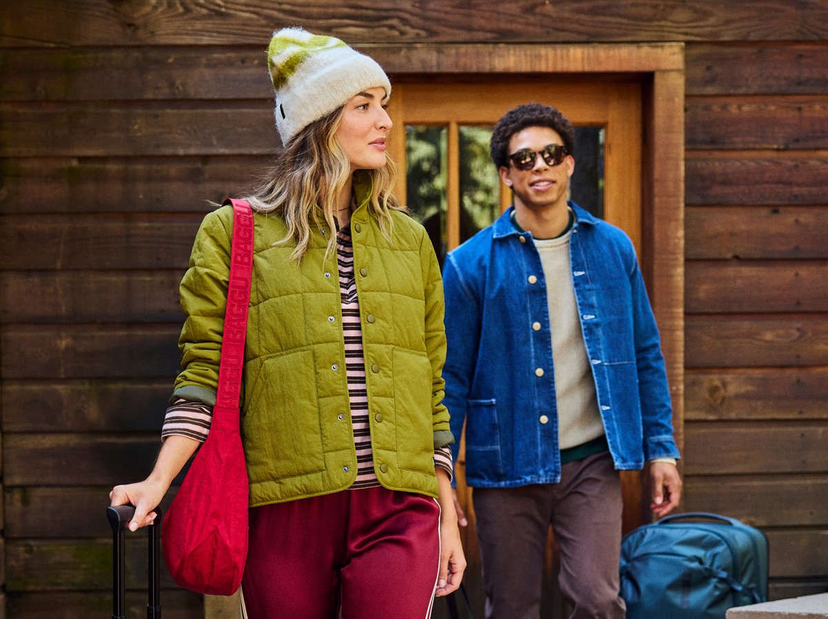 Two people leaving a wood cabin with luggage, wearing casual cold-weather layers including a quilted vest, beanie, and denim jacket.