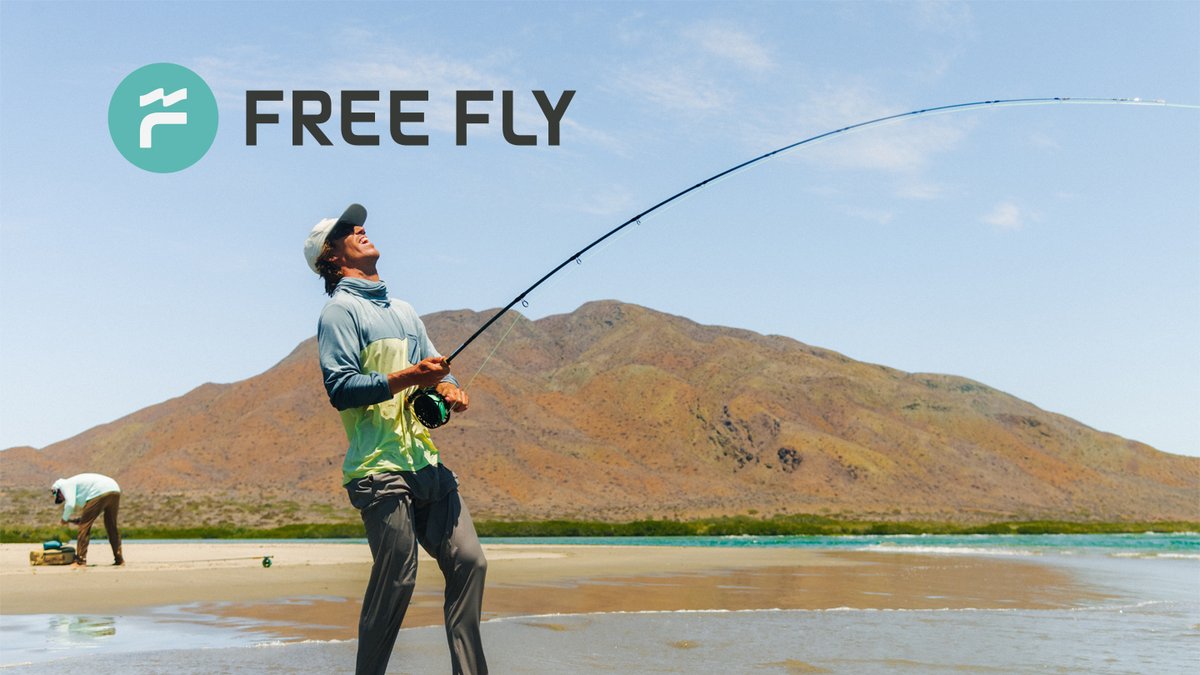 A fly fisherman casts his line in shallow turquoise waters with desert mountains in the background under clear skies.