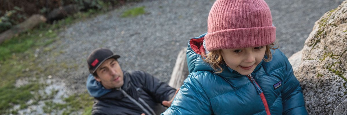 Young child wearing a teal down jacket and pink beanie climbing rocks outdoors while an adult supervises from below on a rocky terrain.