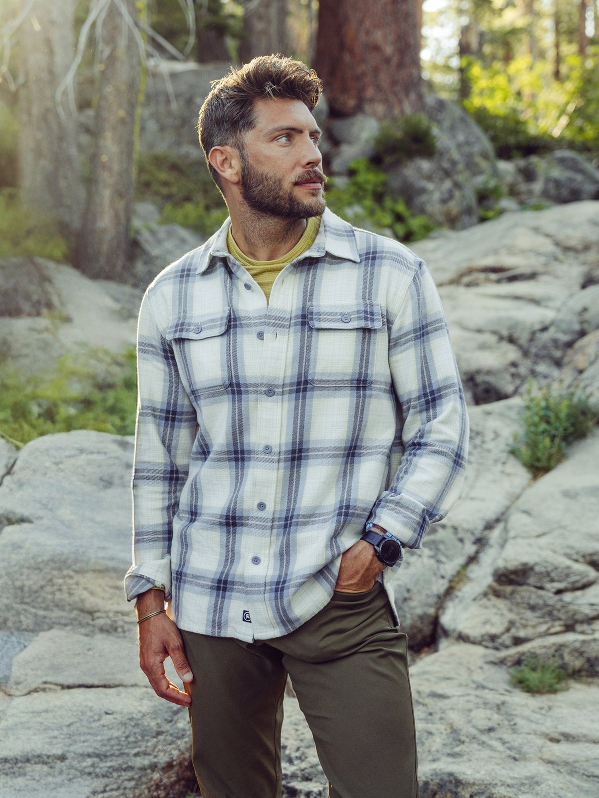 A man stands outdoors on rocky terrain wearing a white plaid button-up shirt over a yellow undershirt and olive green pants, surrounded by trees and forest scenery.