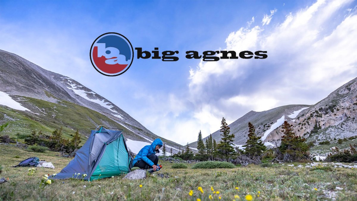 A camper in a blue down jacket prepares gear next to a small green tent in a vast mountain meadow under the Big Agnes logo.