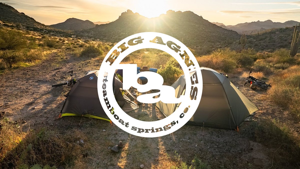 A camper in a blue down jacket prepares gear next to a small green tent in a vast mountain meadow under the Big Agnes logo.