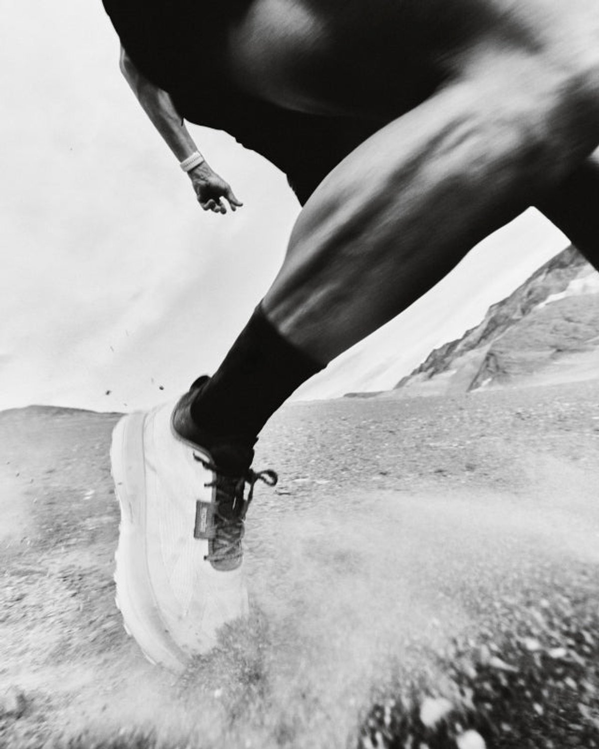 Black-and-white ground-level shot of a Norda trail shoe striking a rocky alpine surface, with motion-blurred legs above and a mountain ridgeline behind.