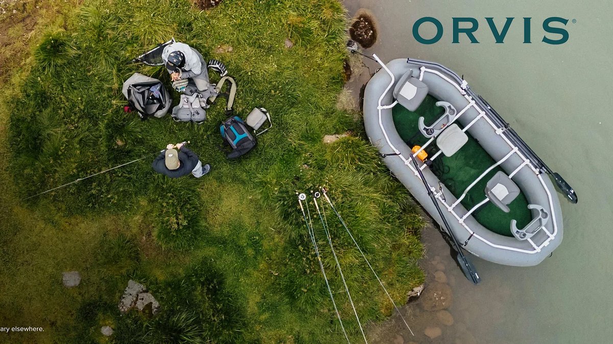 Overhead view of an angler in waders standing on mossy ground next to an Orvis inflatable boat moored in shallow water with fishing gear and backpacks nearby.