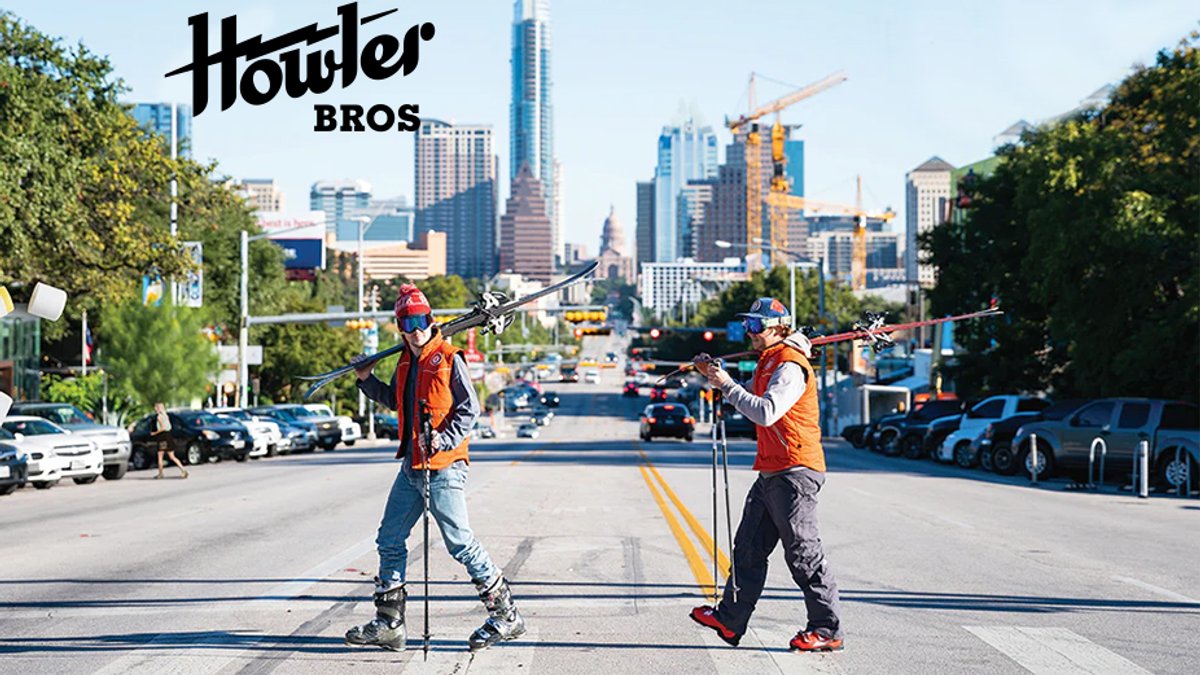 Two men wearing orange vests and ski gear walk across a city street carrying skis, with the Howler Bros logo in the corner.