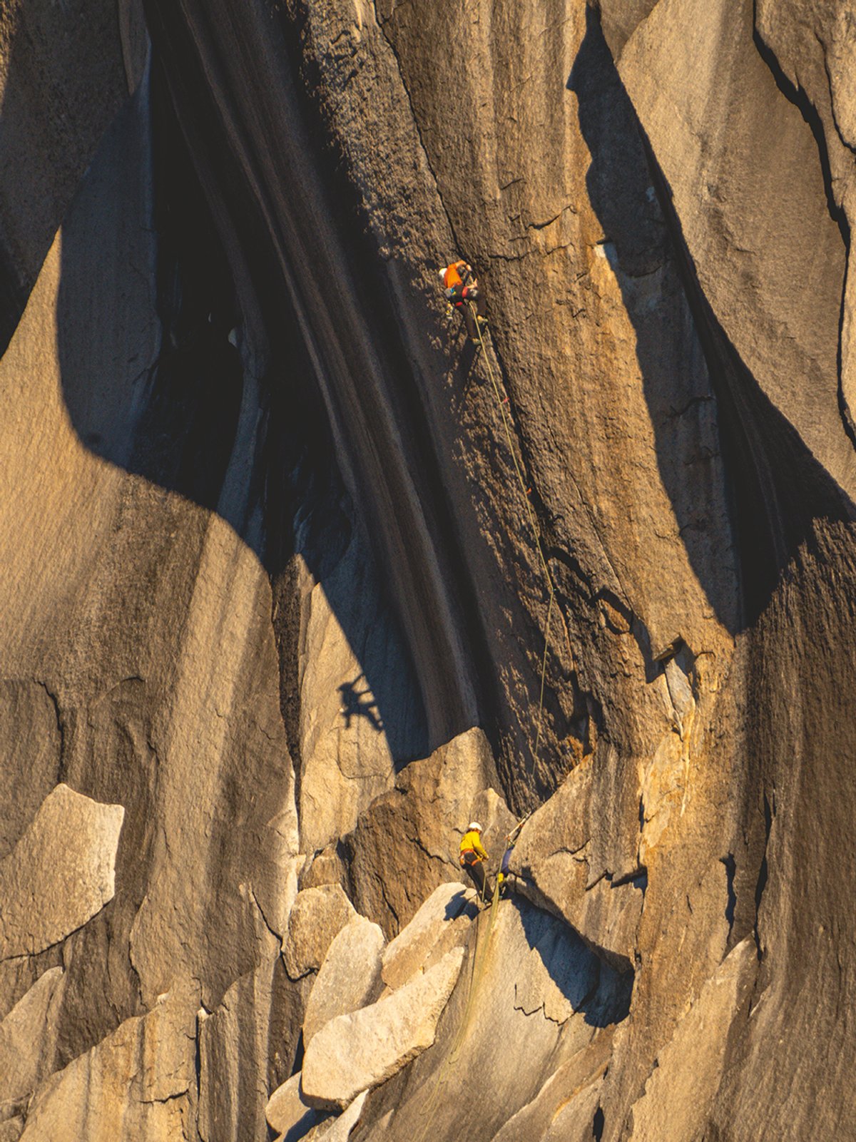 Two rock climbers ascending massive granite multipitch wall above valley floor in golden evening light.