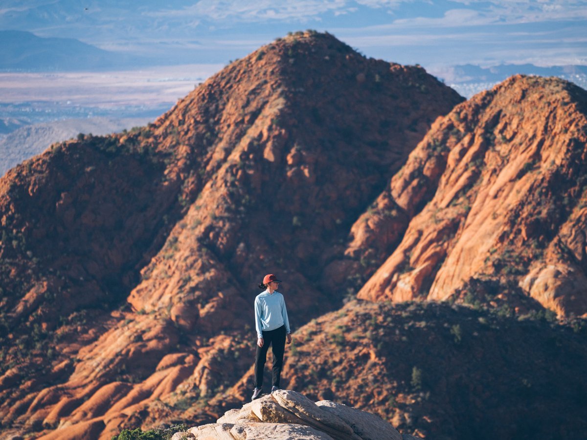 Person in a light blue long-sleeve shirt and red cap standing on a rock outcrop above red sandstone ridgelines, with a valley and distant mountains in the background.