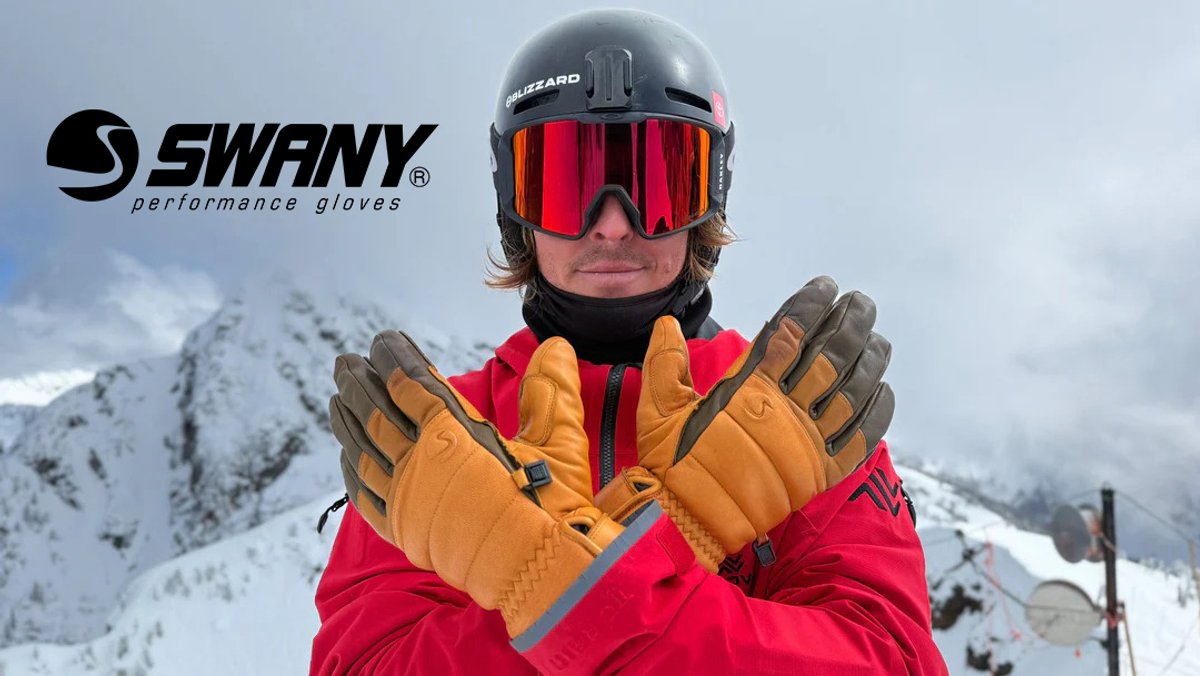 Skier wearing orange and brown Swany performance gloves with red jacket and red-lens goggles standing in snowy mountain terrain.