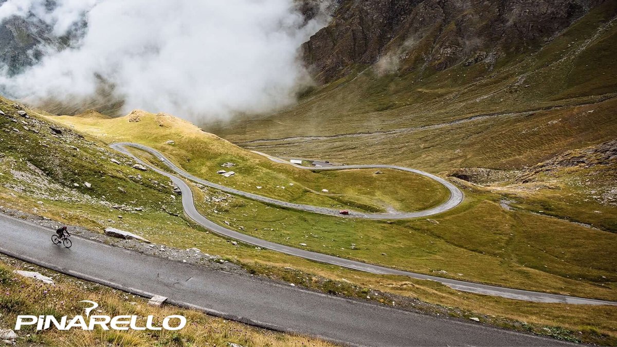 Cyclist riding down winding mountain road through grassy hills with cloudy valley in the background.
