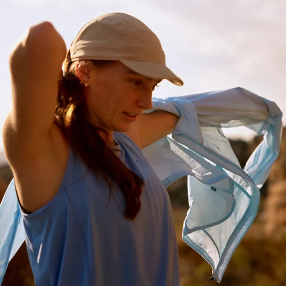 Woman wearing a light blue cap and matching cooling towel draped over her shoulder in bright outdoor sunlight, demonstrating sun protection and athletic gear usage.