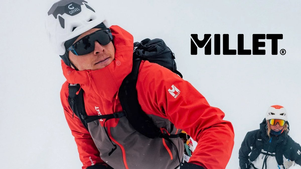 Man wearing Millet red ski jacket and white helmet with sunglasses skiing in snowy conditions with another skier visible in background.