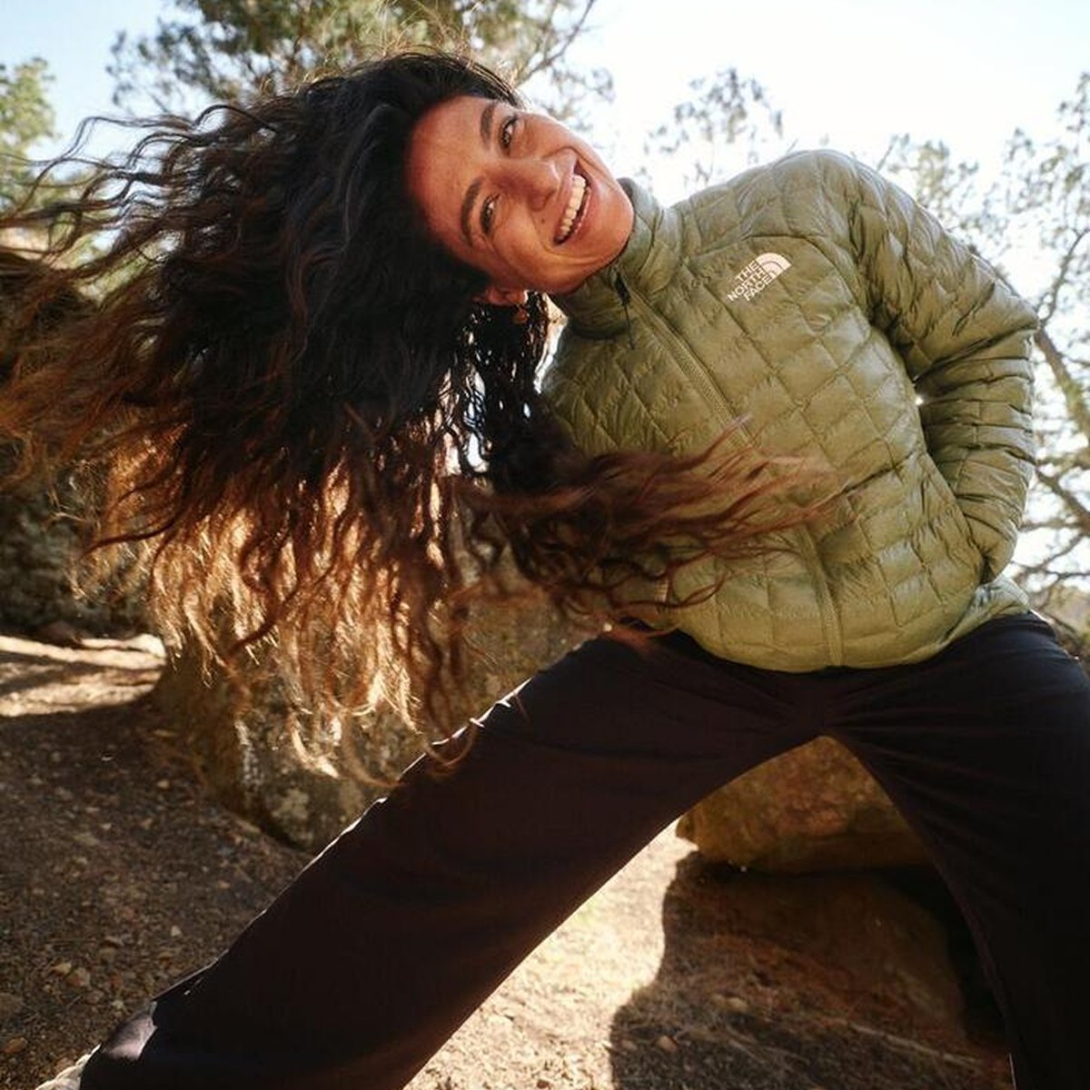 A smiling woman in a green puffer jacket tosses her long, dark, curly hair back while standing outdoors.