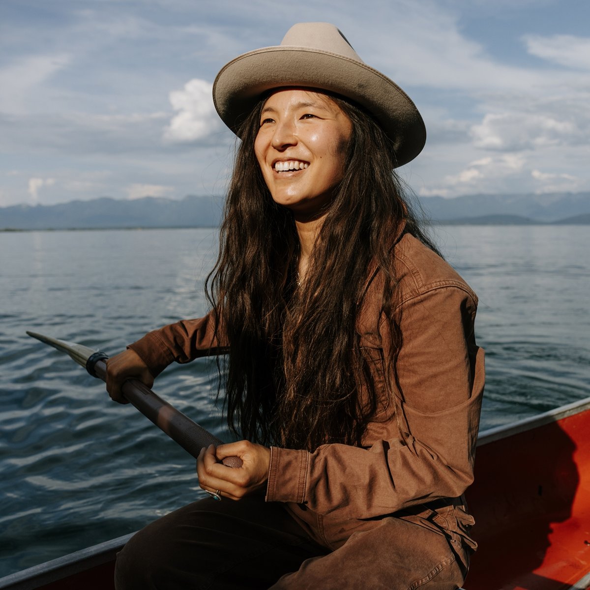 A woman in a wide-brimmed hat and brown jumpsuit paddles a red canoe.