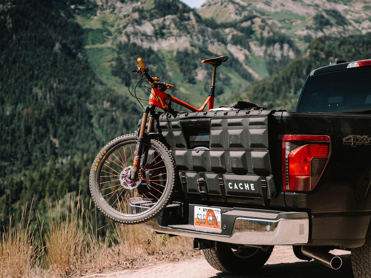 Orange enduro bike secured over a black CACHE Basecamp 3.0 tailgate pad on a truck, mountain hillside in the background.