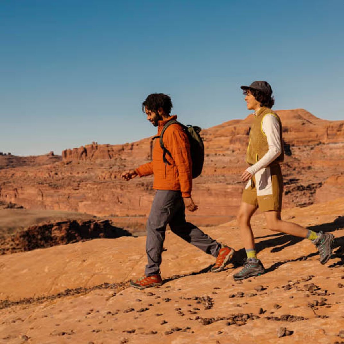 Two hikers walk across a red rock plateau wearing Merrell hiking shoes, with canyon formations visible in the background under a clear blue sky.