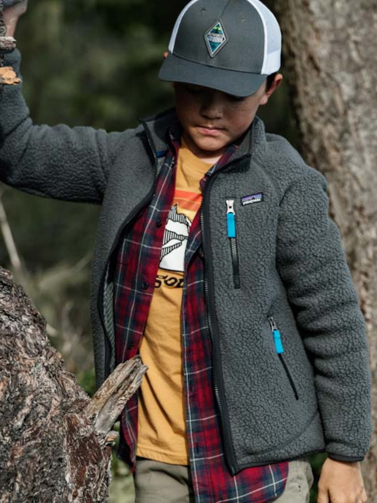 A boy wearing a gray Patagonia fleece jacket over a plaid shirt climbs a tree.