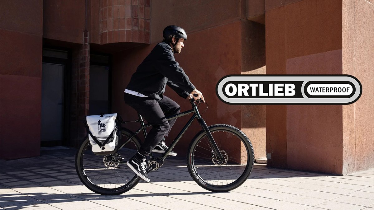 A cyclist wearing a black helmet and jacket rides a gravel bike equipped with a waterproof Ortlieb pannier bag through an urban brick courtyard in daylight.