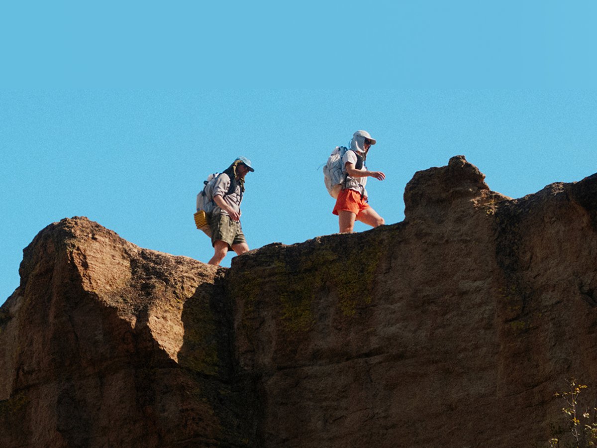Two hikers traversing a sun-exposed sandstone ridgeline with loaded packs against a clear blue sky.