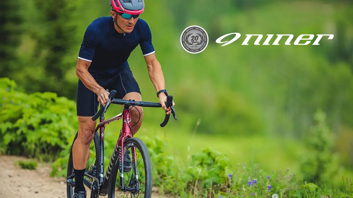 A cyclist in navy athletic wear and a pink helmet rides a red road bike on a scenic country trail surrounded by lush green vegetation.