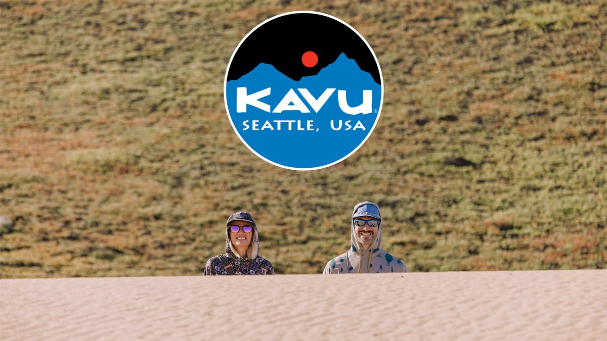 Two people in hoodies stand behind a sand dune with a large circular KAVU logo centered above them.