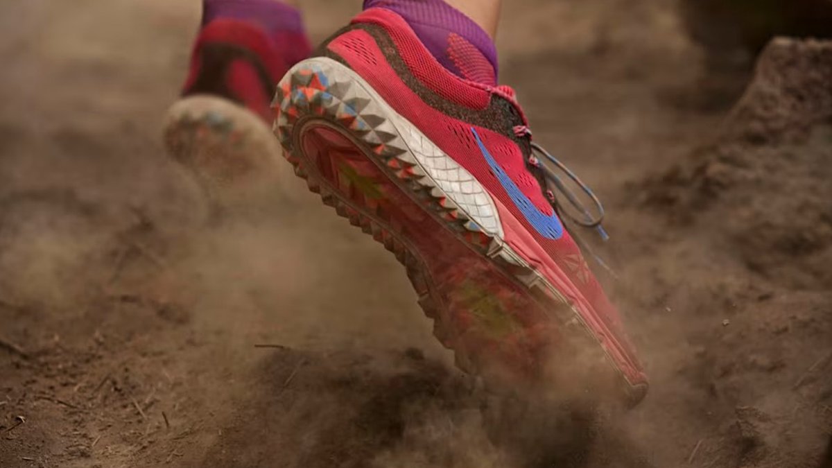 A close-up action shot of a bright pink Nike trail running shoe kicking up dust on a dirt path.