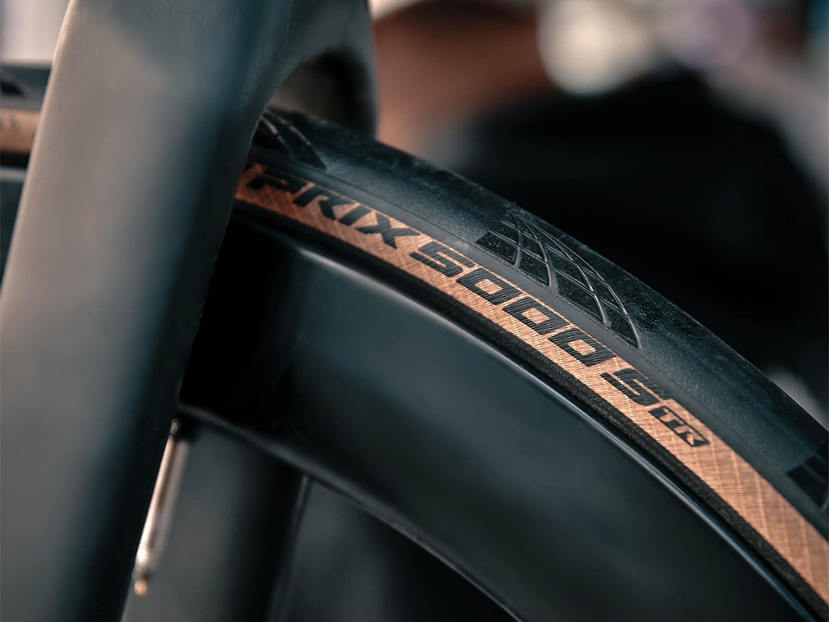 Close-up of a Continental Grand Prix 5000 S TR tire with tan sidewall on a deep-section carbon wheel, shot through a dark carbon fork in low light.