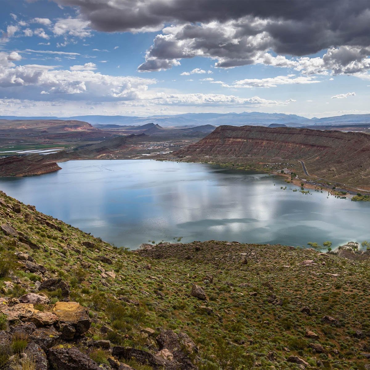 Aerial view of Quail Creek Reservoir nestled below a red sandstone mesa, with desert scrub in the foreground and mountains receding to the horizon under a partly cloudy sky.