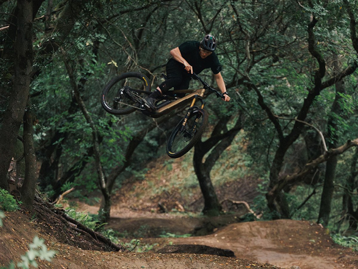 Mountain biker wearing MTB helmet and shorts sending jump on full-suspension mountain bike through wooded forest singletrack trail