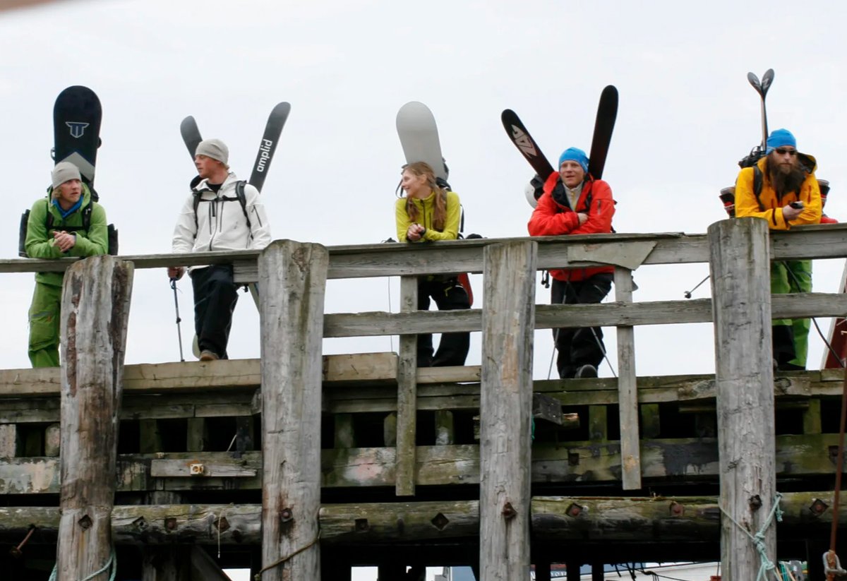 Group of five skiers and snowboarders standing on a wooden dock holding their equipment while wearing colorful winter jackets and beanies against a gray sky.